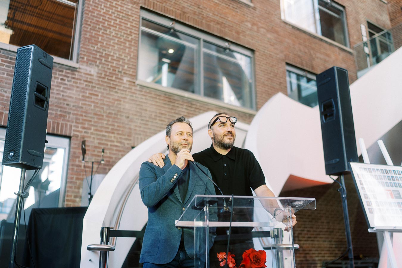 Two men stand at a clear podium with microphones in a modern atrium space at Ricarda’s in Toronto, speaking at the Toronto Prep Forever Launch Party.