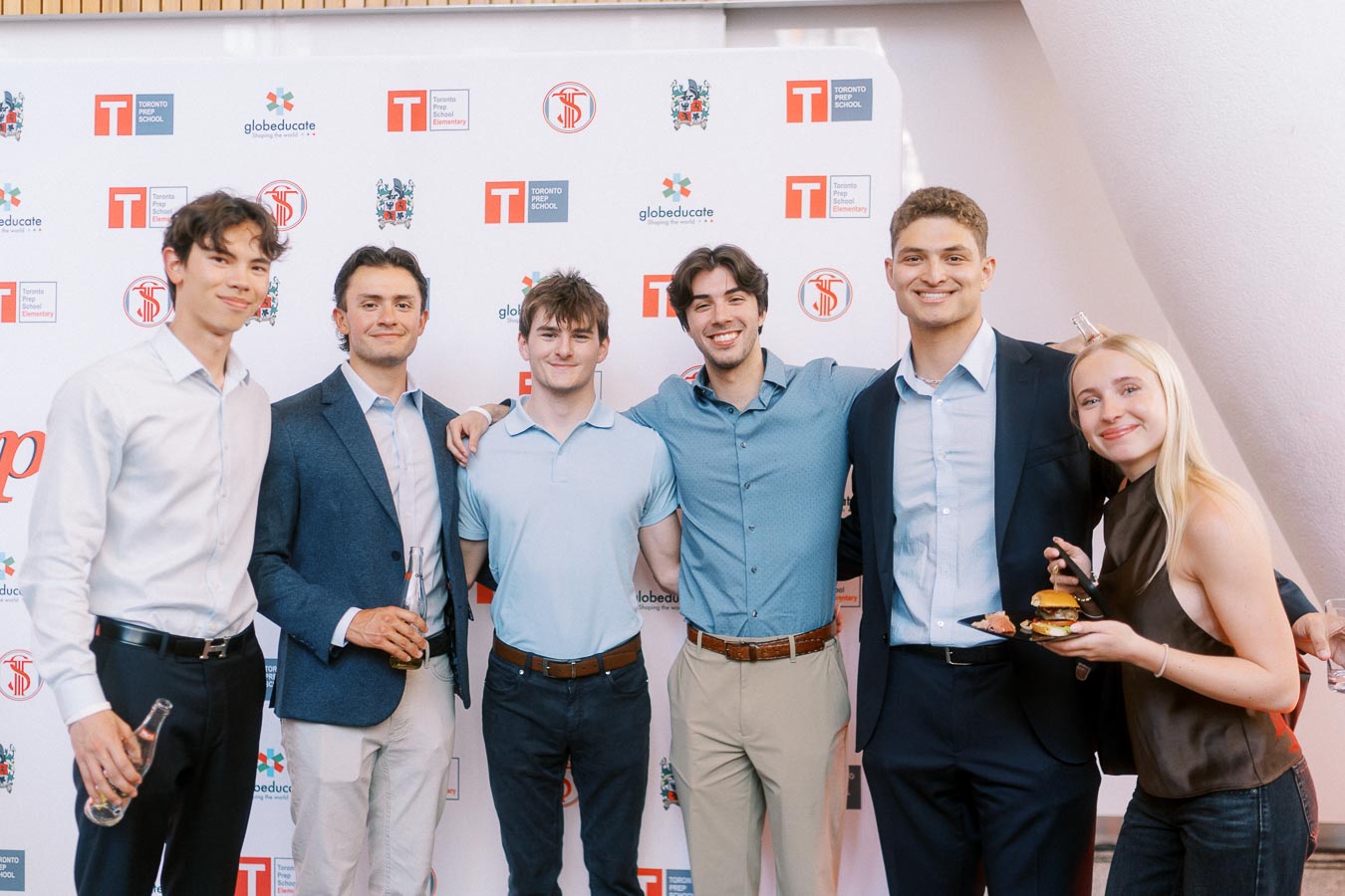 Six young adults stand smiling in front of a Toronto Prep School branded backdrop at Ricarda’s in Toronto during the Forever Launch Party.