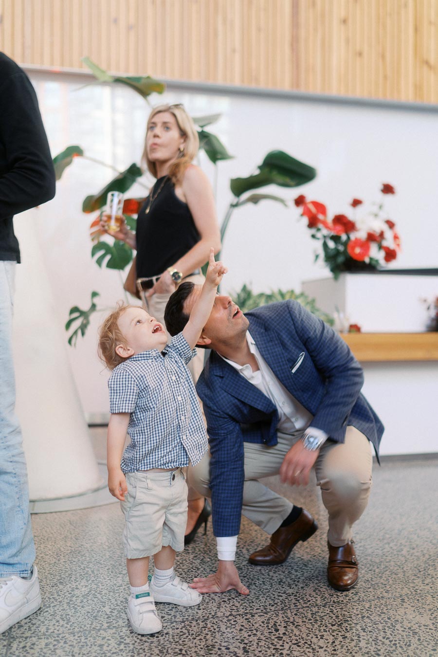 A young child pointing upward next to a crouching adult in a blue blazer at Ricarda’s Atrium during the Toronto Prep Forever Launch Party, with other guests and floral arrangements visible.