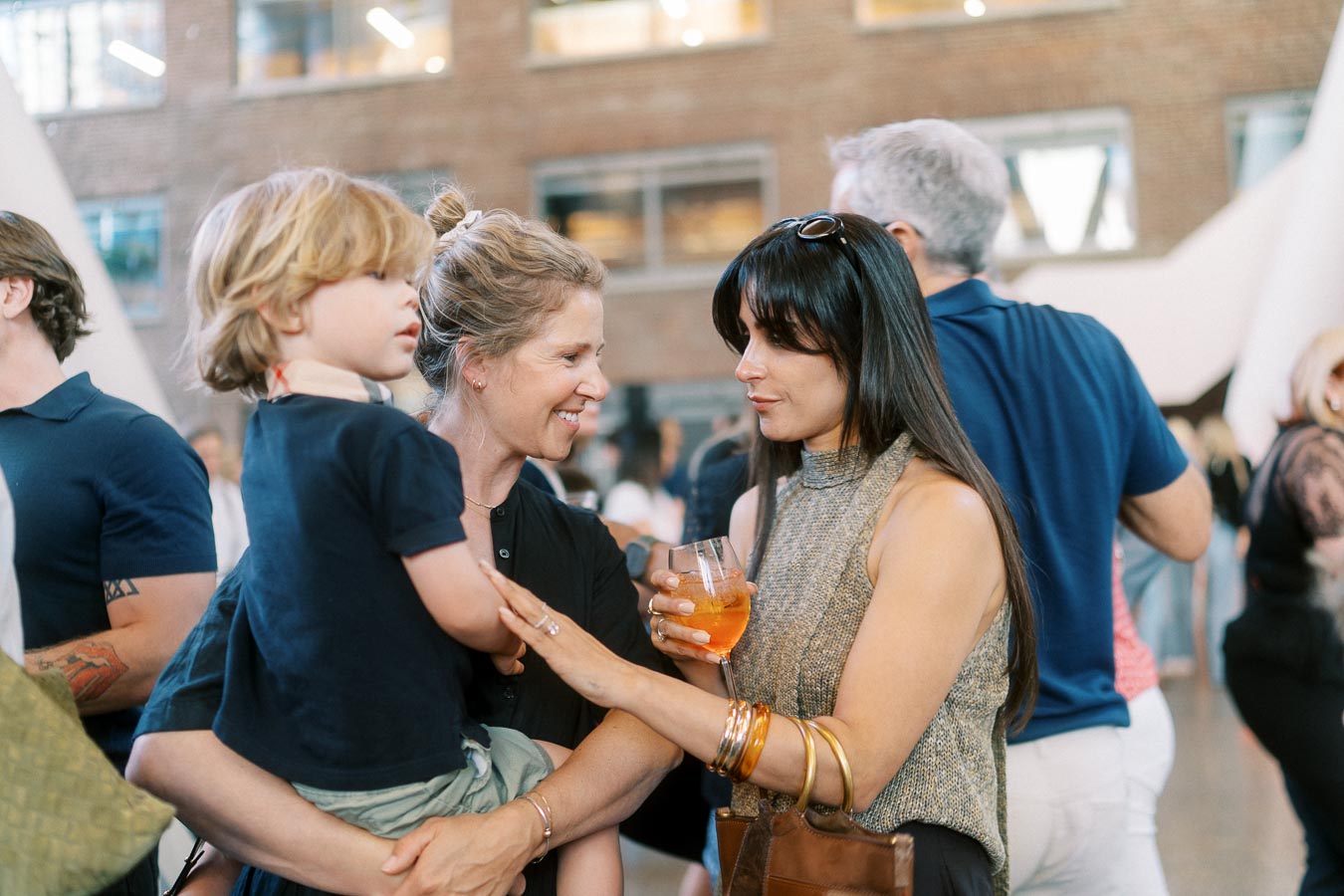 Two women talking and smiling, one holding a child, at the Toronto Prep Forever Launch Party at Ricarda’s in Toronto, with guests mingling in the background.