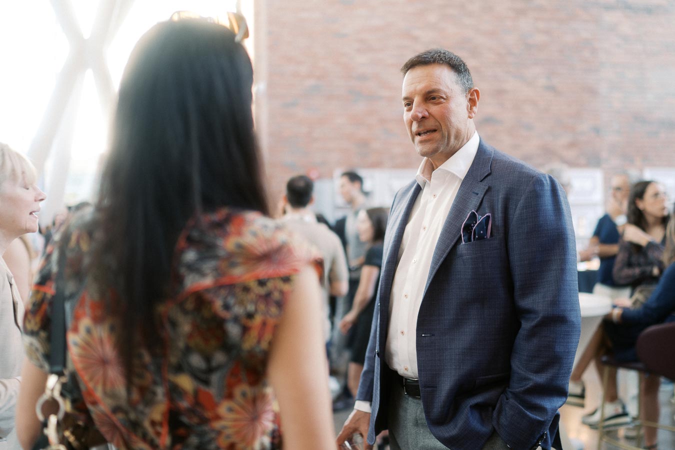 A man in a blue suit converses with a woman in a patterned dress at the Toronto Prep Forever Launch Party at Ricarda’s, surrounded by event guests.