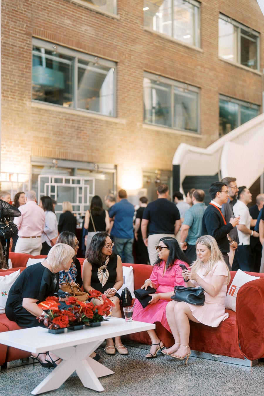 Four women seated on red couches, conversing and using a phone, with a crowd mingling in the background at Ricarda’s Toronto Prep Forever Launch Party.