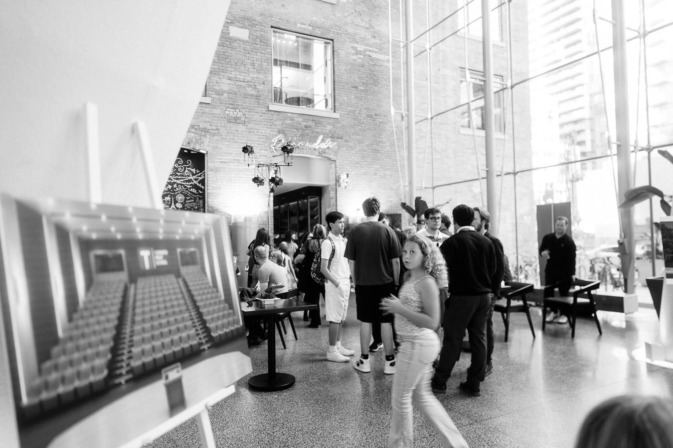 A group of guests gather inside The Atrium at Ricarda’s in Toronto, with a blurred display of a school auditorium rendering in the foreground and modern glass architecture visible.
