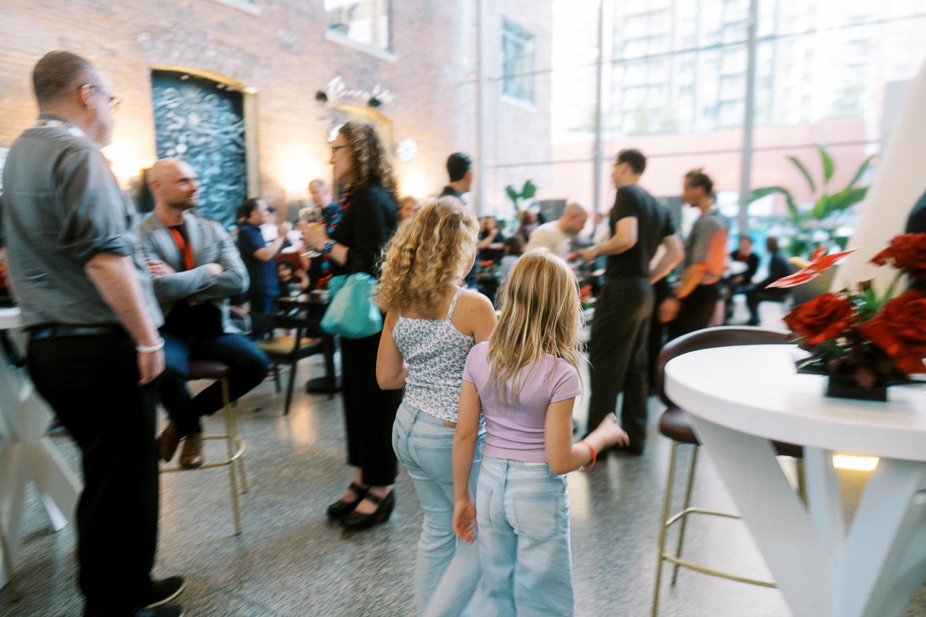 Two children walking among adults at a busy Toronto Prep School launch event in Ricarda’s Atrium, with guests mingling in a modern glass venue.