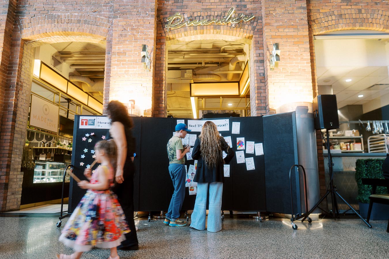 Children and adults participating in an interactive board activity at the Toronto Prep Forever Launch Party held inside Ricarda’s, Toronto, with visible brick walls and event signage.