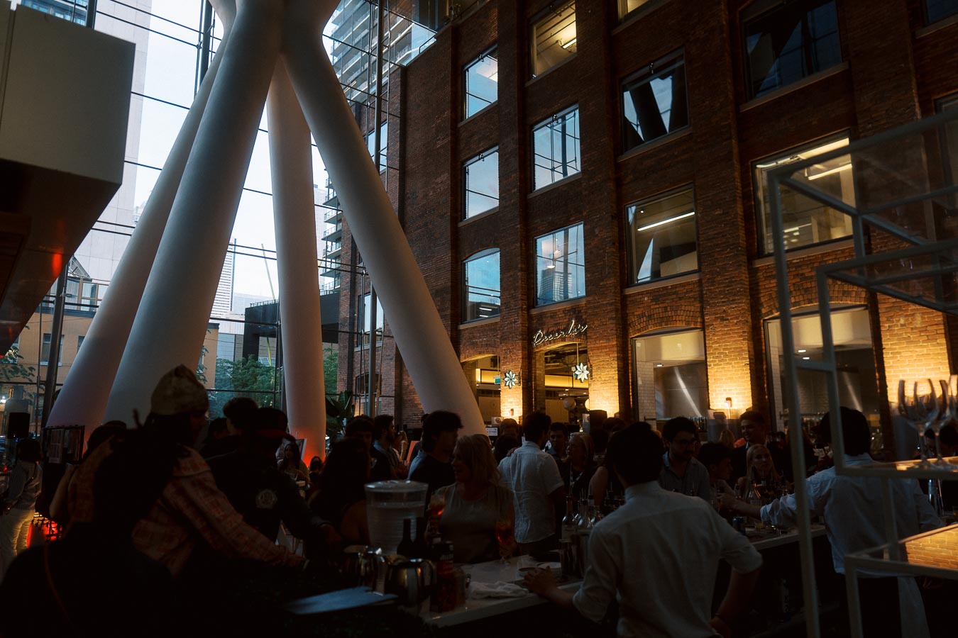 A lively crowd enjoys drinks and conversation at the bar in The Atrium at Ricarda’s, Toronto, with exposed brick walls and modern glass architecture visible in the background.