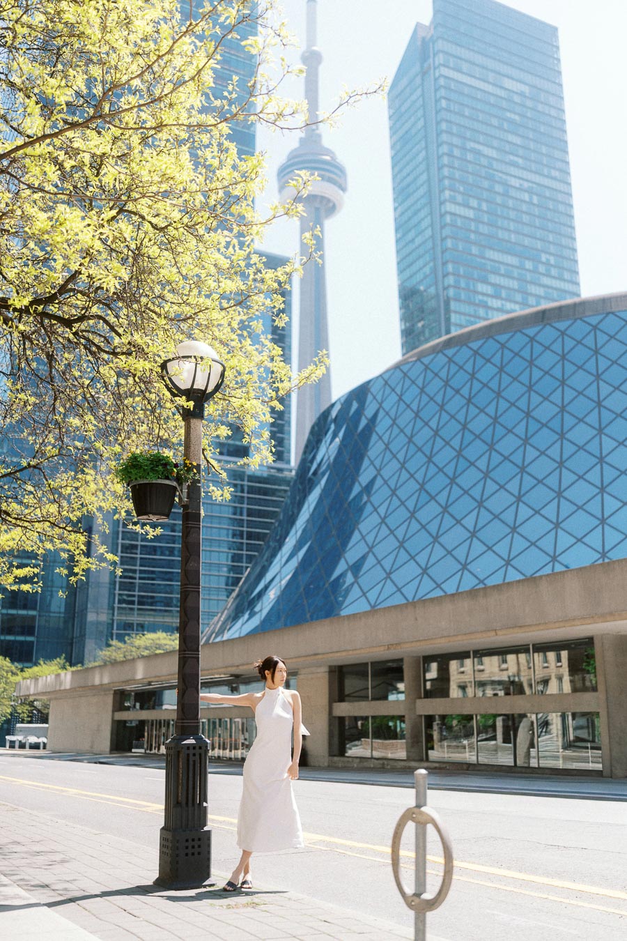 Woman in a white dress leaning on a lamppost on a sunny Toronto street, CN Tower and modern glass building behind her.