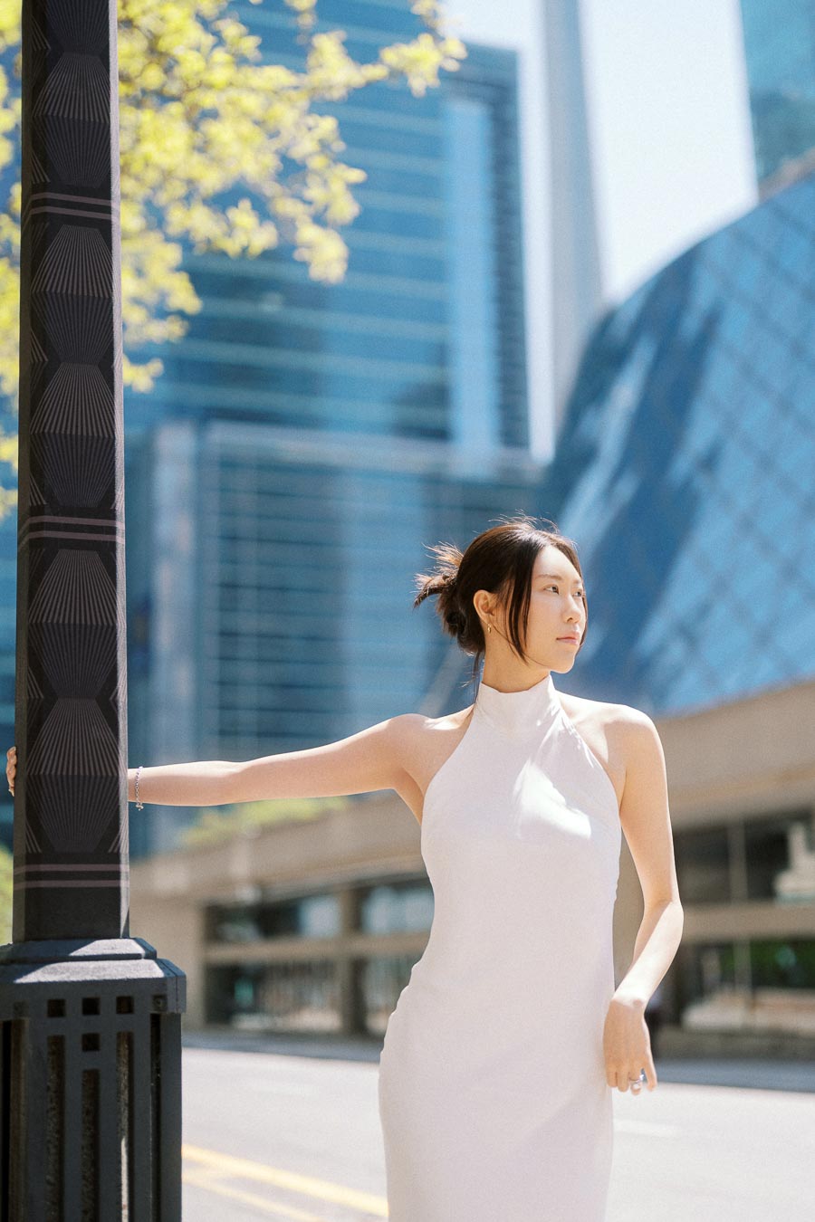 Woman in a white dress leaning on a streetlamp in downtown Toronto with modern glass buildings in the background.