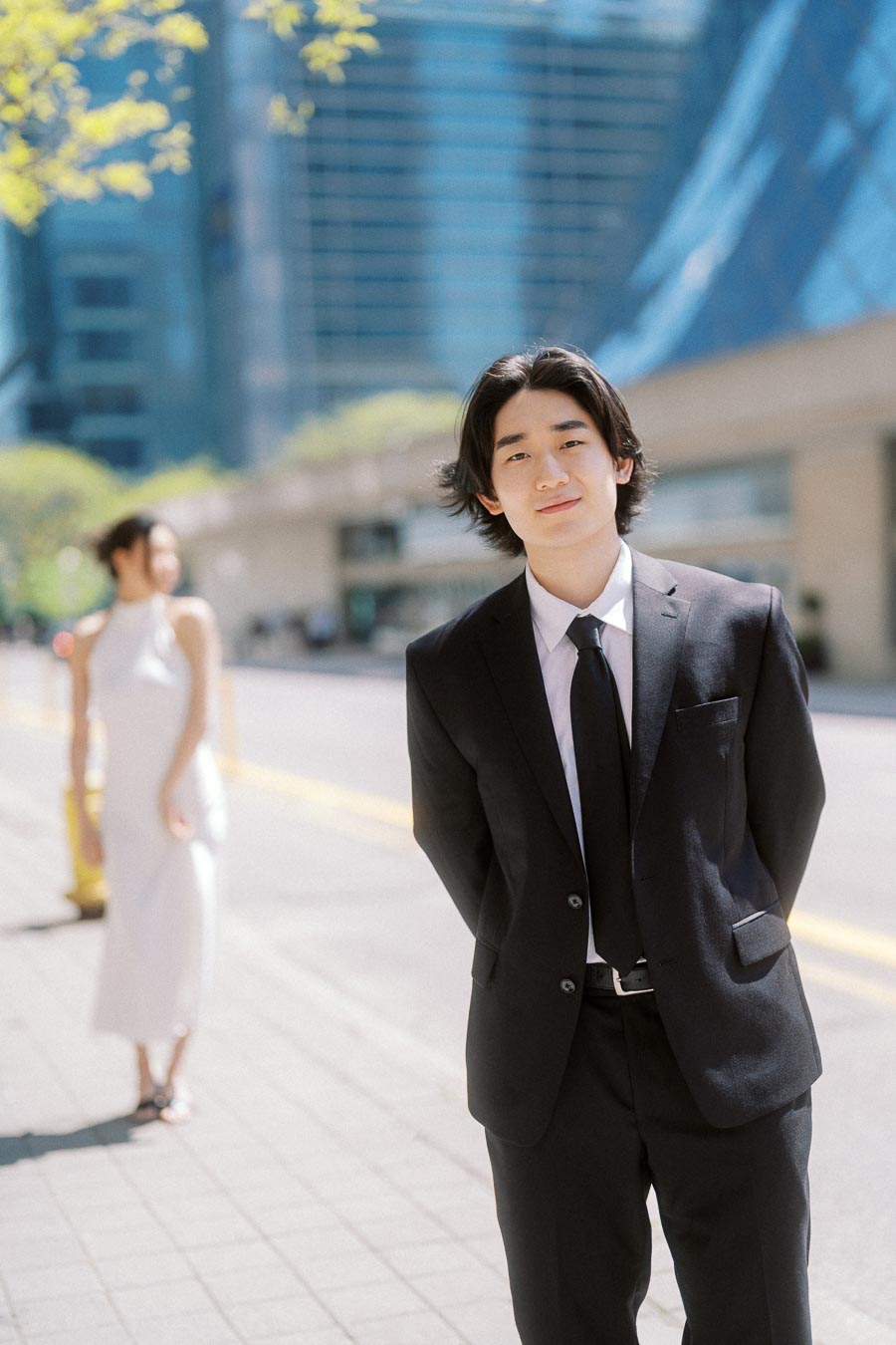 Person in black suit posing in focus on a bright city sidewalk, with their partner in a white dress blurred behind them.