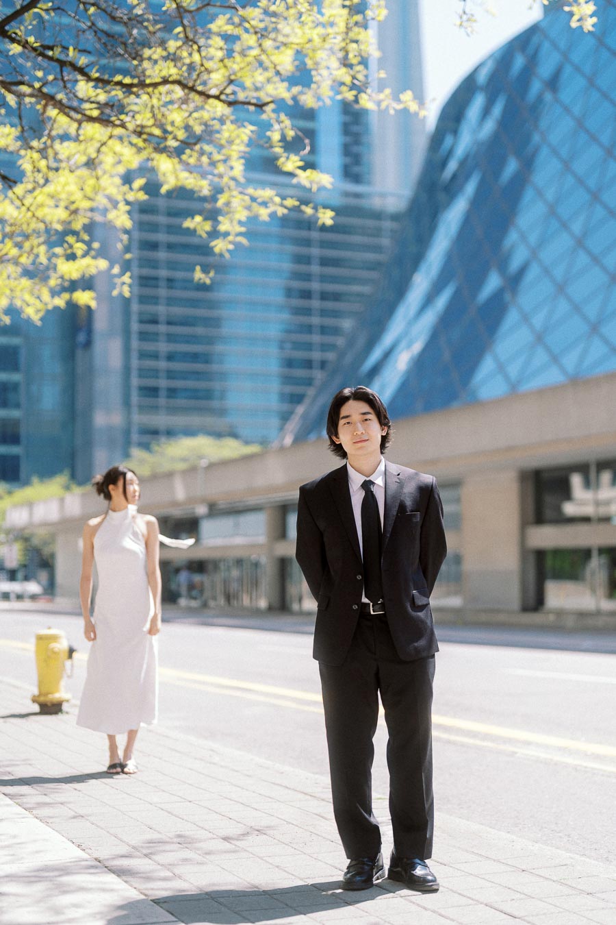 Person in a black suit stands in focus on a city sidewalk while their partner in a white dress walks behind, with glass buildings and spring tree branches overhead.