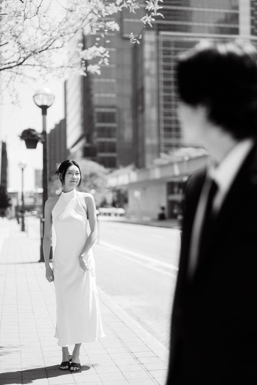 Person in a white dress stands on a city sidewalk while another in a suit looks on, downtown Toronto skyline in background.