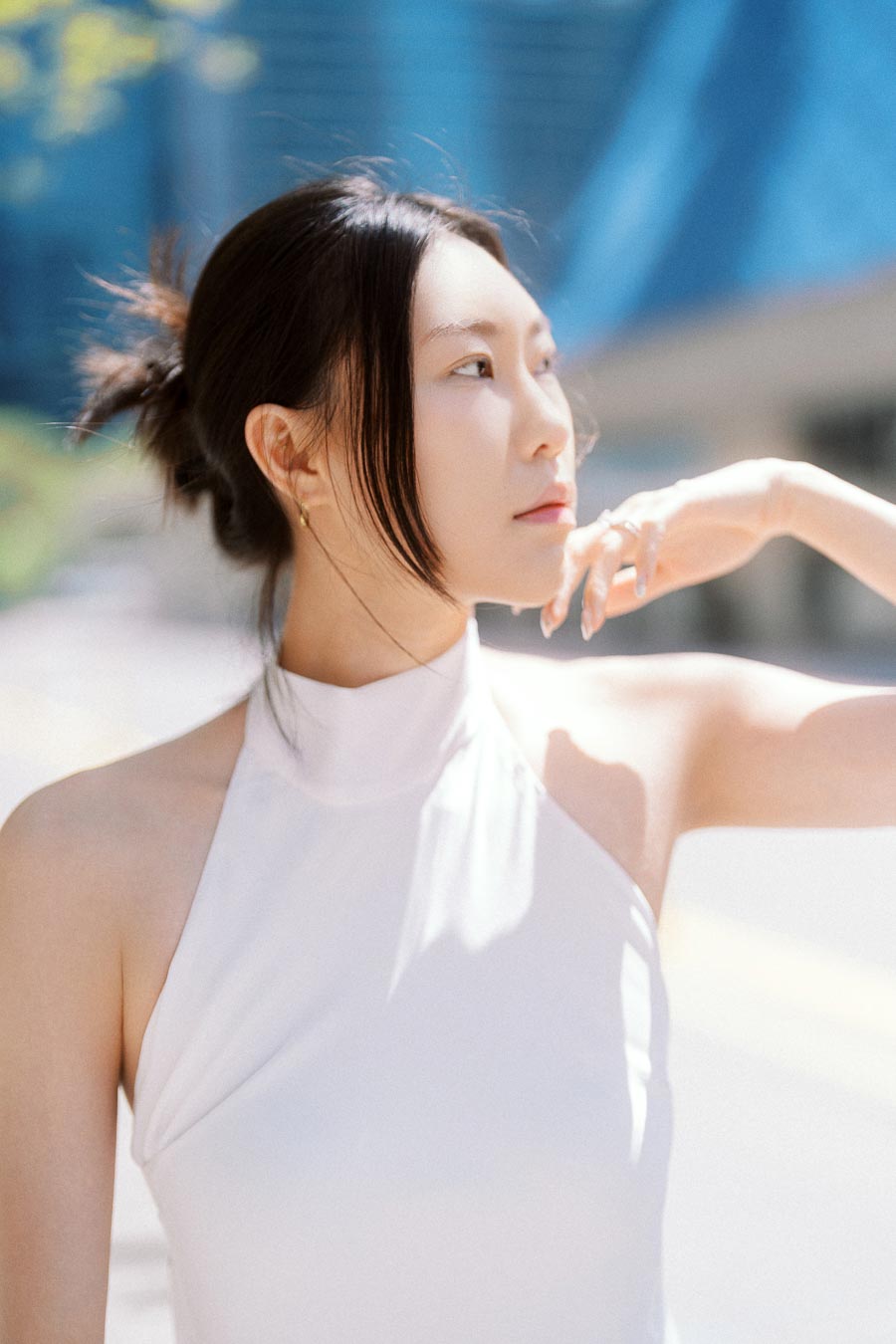 Woman in a white halter dress posing in sunlight with blurred urban architecture behind her.