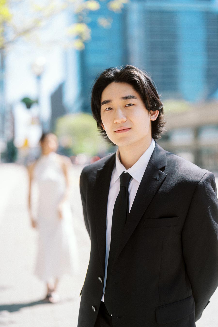 Man in a black suit stands in focus on a city sidewalk, with a woman in a white dress softly blurred behind him and tall blue buildings in the background.