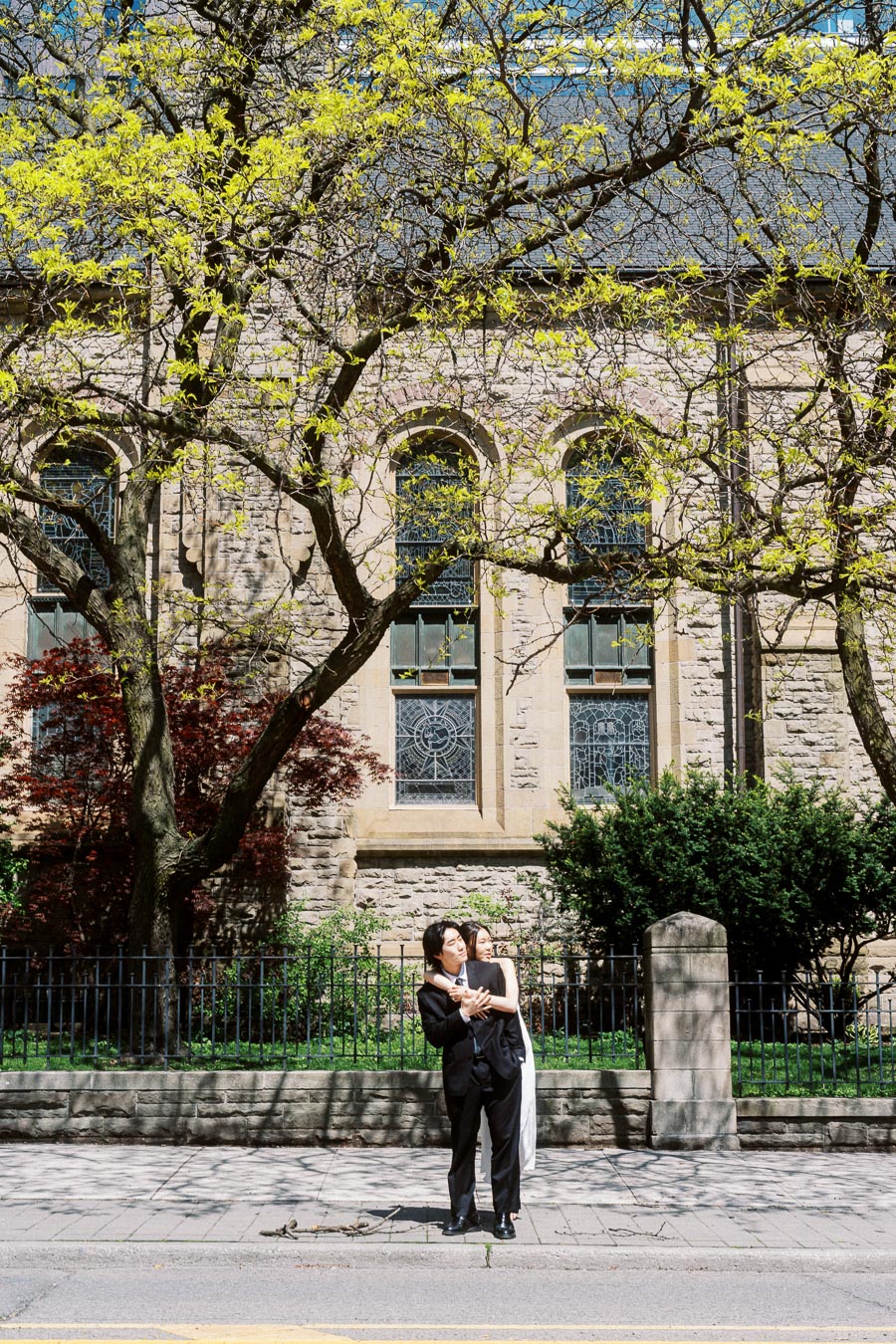 Couple embraces on sidewalk in front of historic stone church with spring trees and stained glass windows.