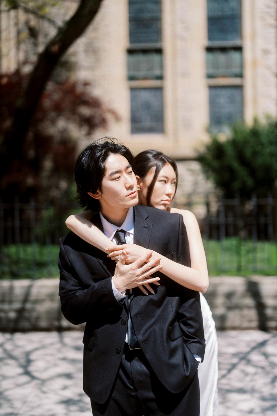 Couple embracing outdoors, one in a black suit and the other in a white dress, with a historic stone church and greenery behind them.