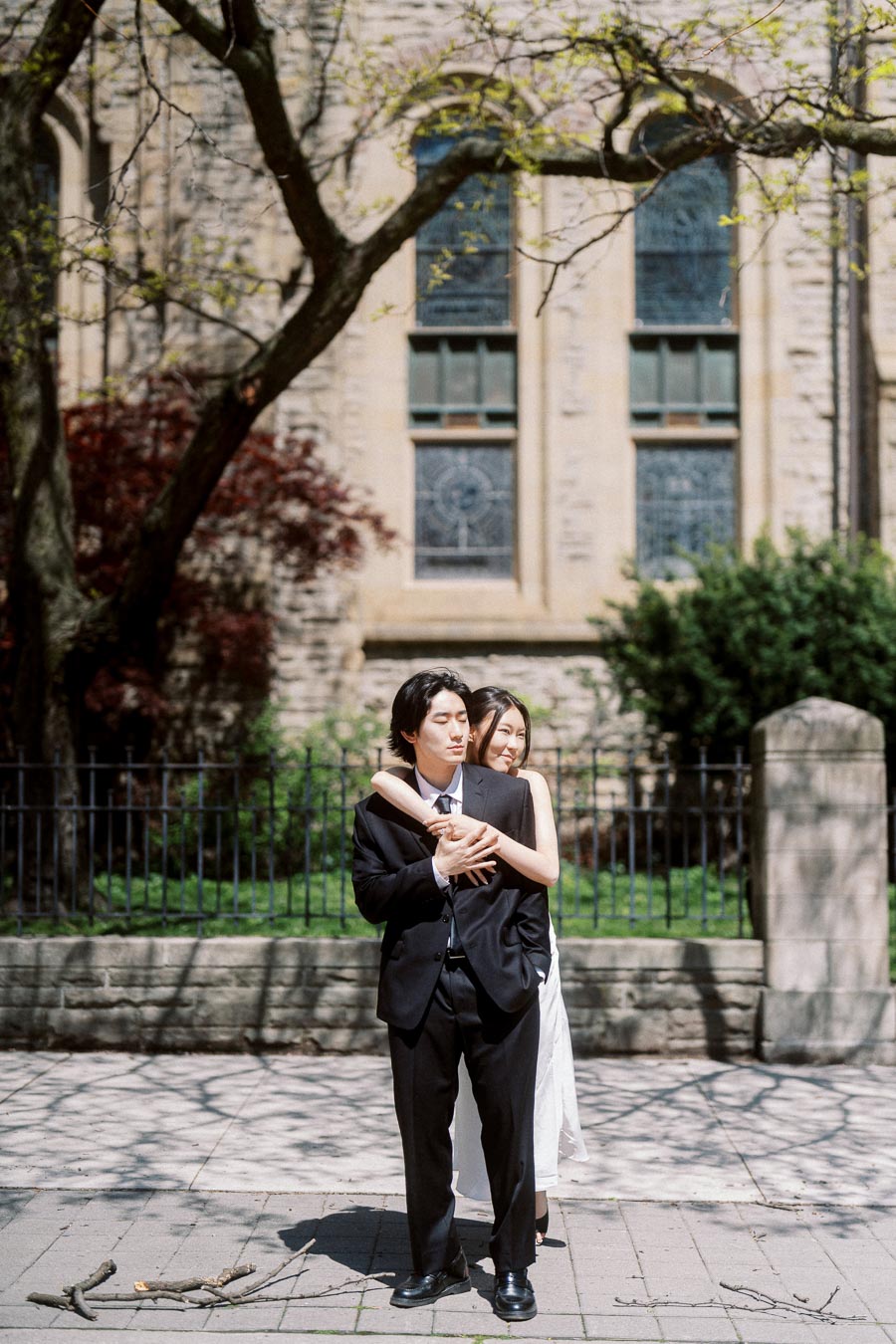 Couple embracing in front of old stone church with arched windows and spring trees in downtown Toronto.