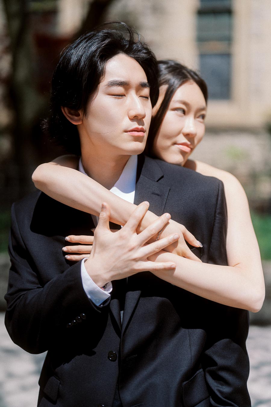 Couple embracing closely outdoors, Jun with eyes closed and Alex smiling softly behind him, both dressed elegantly, in bright natural light.