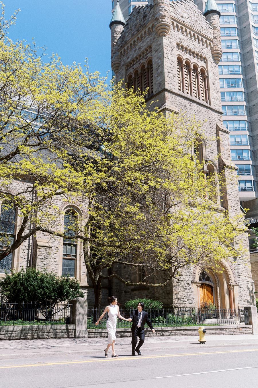 Couple holding hands and walking across the street in front of St. Andrew's Presbyterian Church, with bright spring trees and historic stone architecture in downtown Toronto.