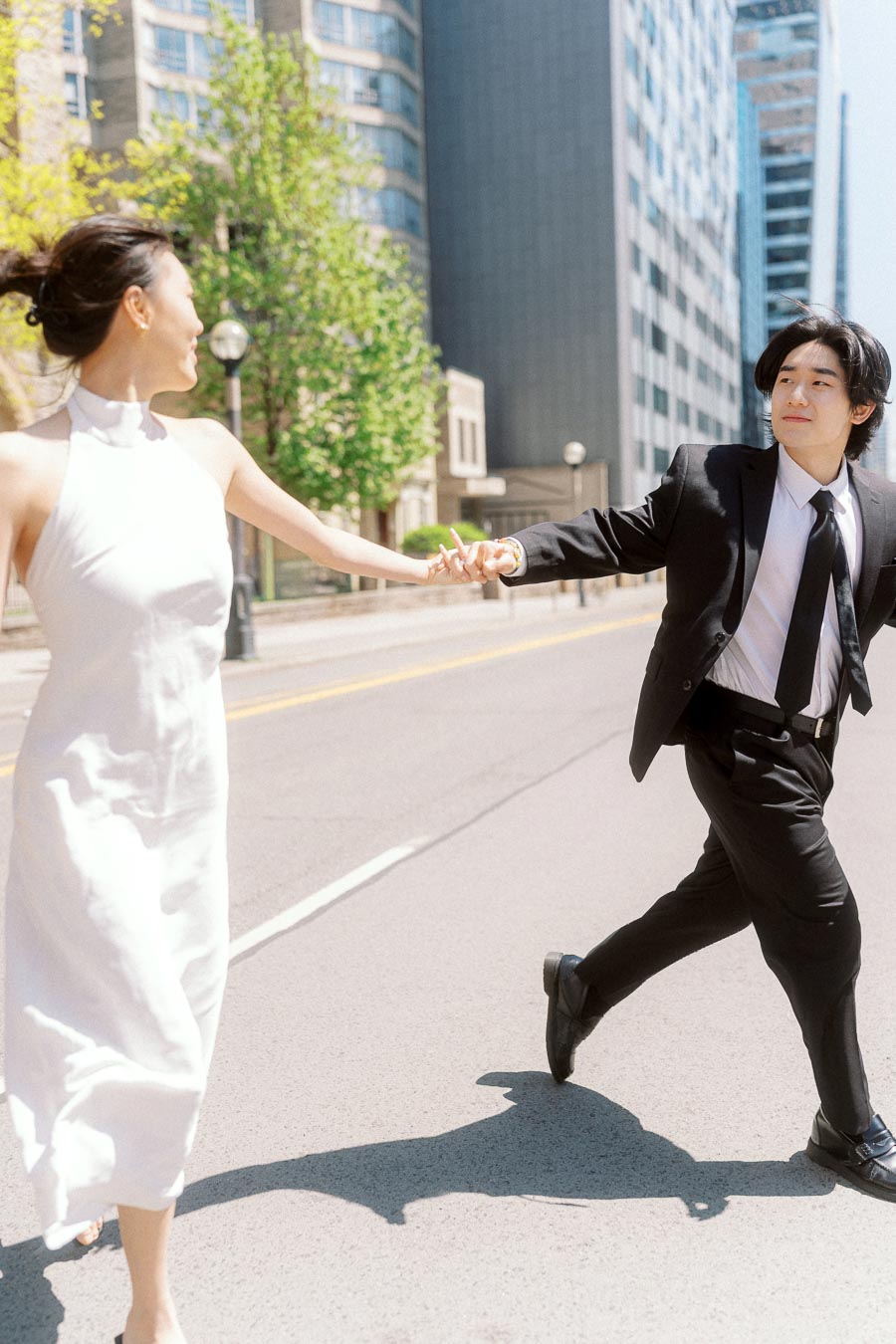 A couple in formal outfits holding hands and crossing an empty city street on a sunny day, surrounded by tall downtown Toronto buildings.