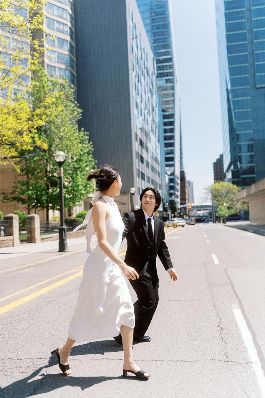 Couple holding hands and crossing a bright city street, surrounded by tall glass buildings and trees in downtown Toronto.
