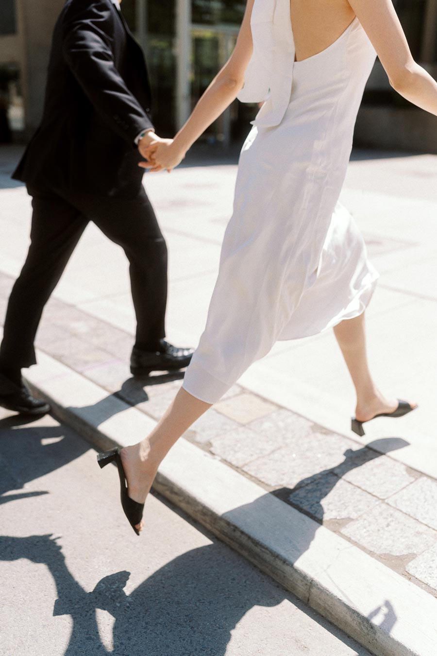 A couple in formal attire holding hands, stepping up onto a city sidewalk in bright daylight.