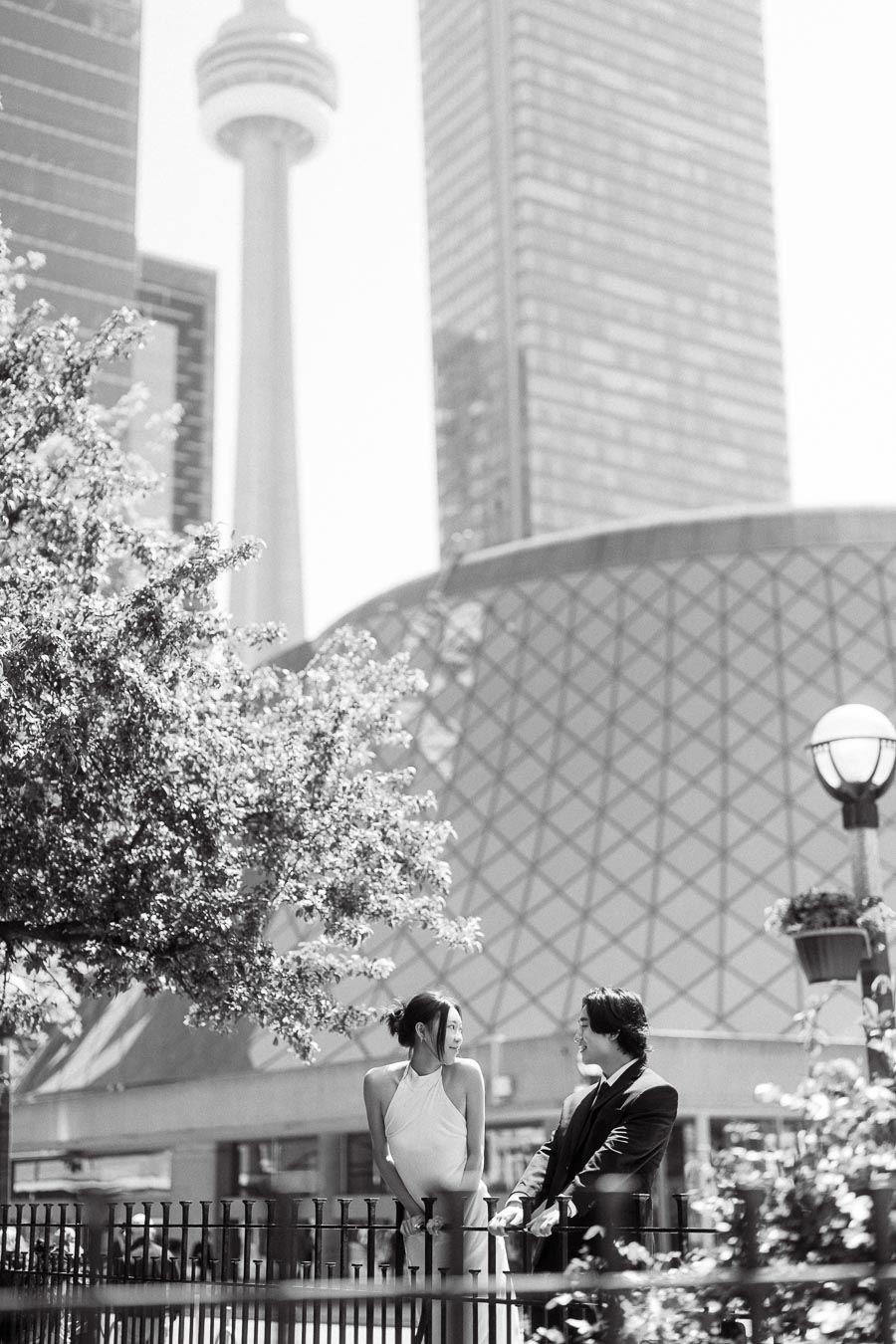 Couple dressed elegantly, smiling at each other near a fence with the CN Tower and downtown Toronto buildings in the background.