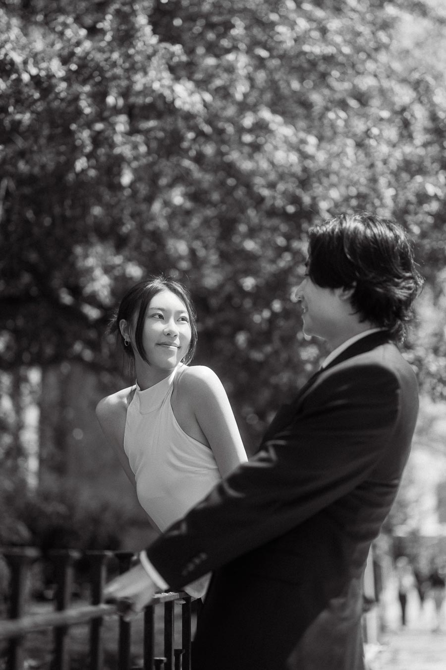 Couple smiling at each other outdoors, woman in white dress and man in dark suit, sunlit trees in background.