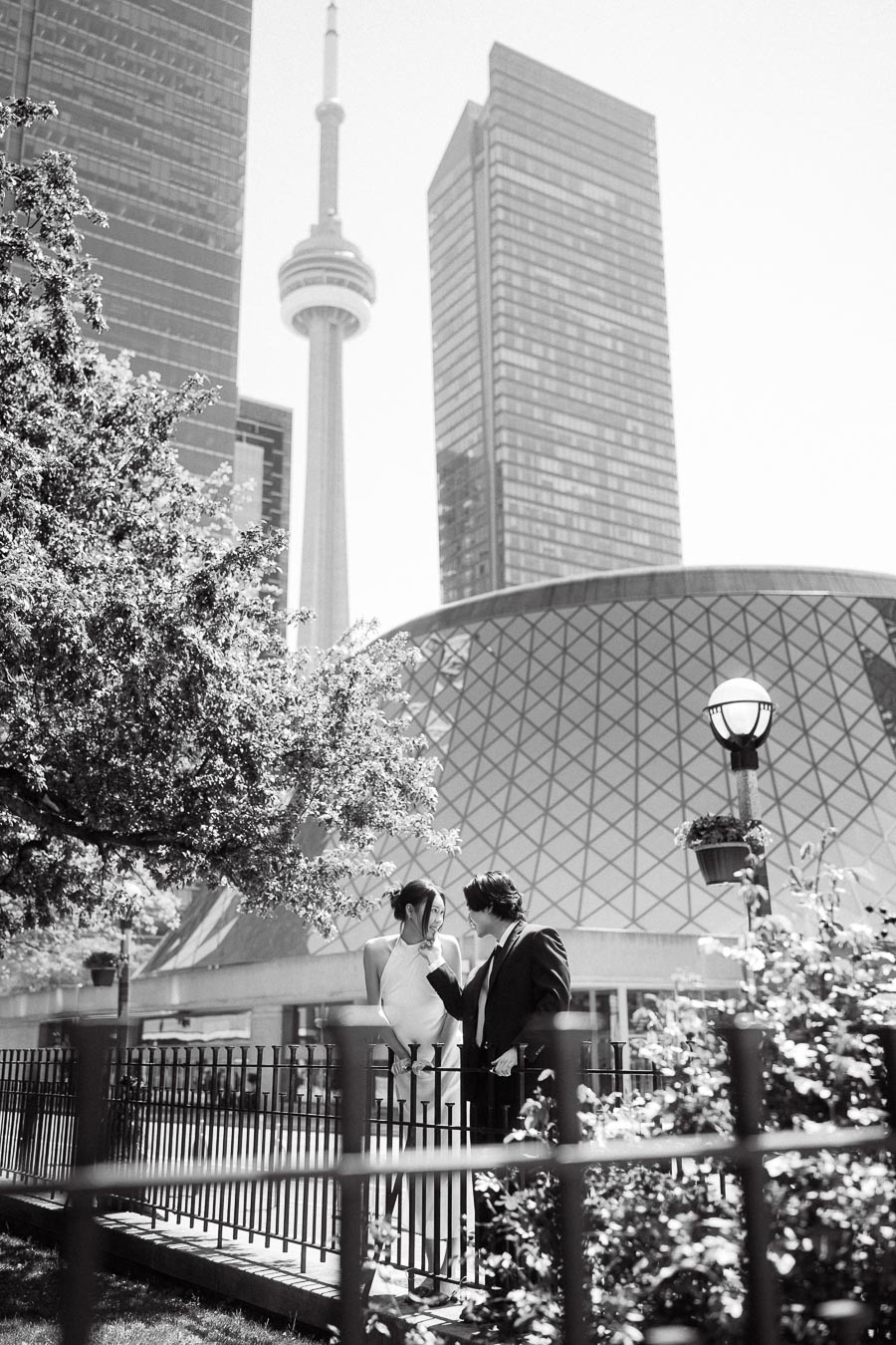 Couple standing close by a garden fence with the CN Tower and city buildings in the background, sharing a romantic moment.
