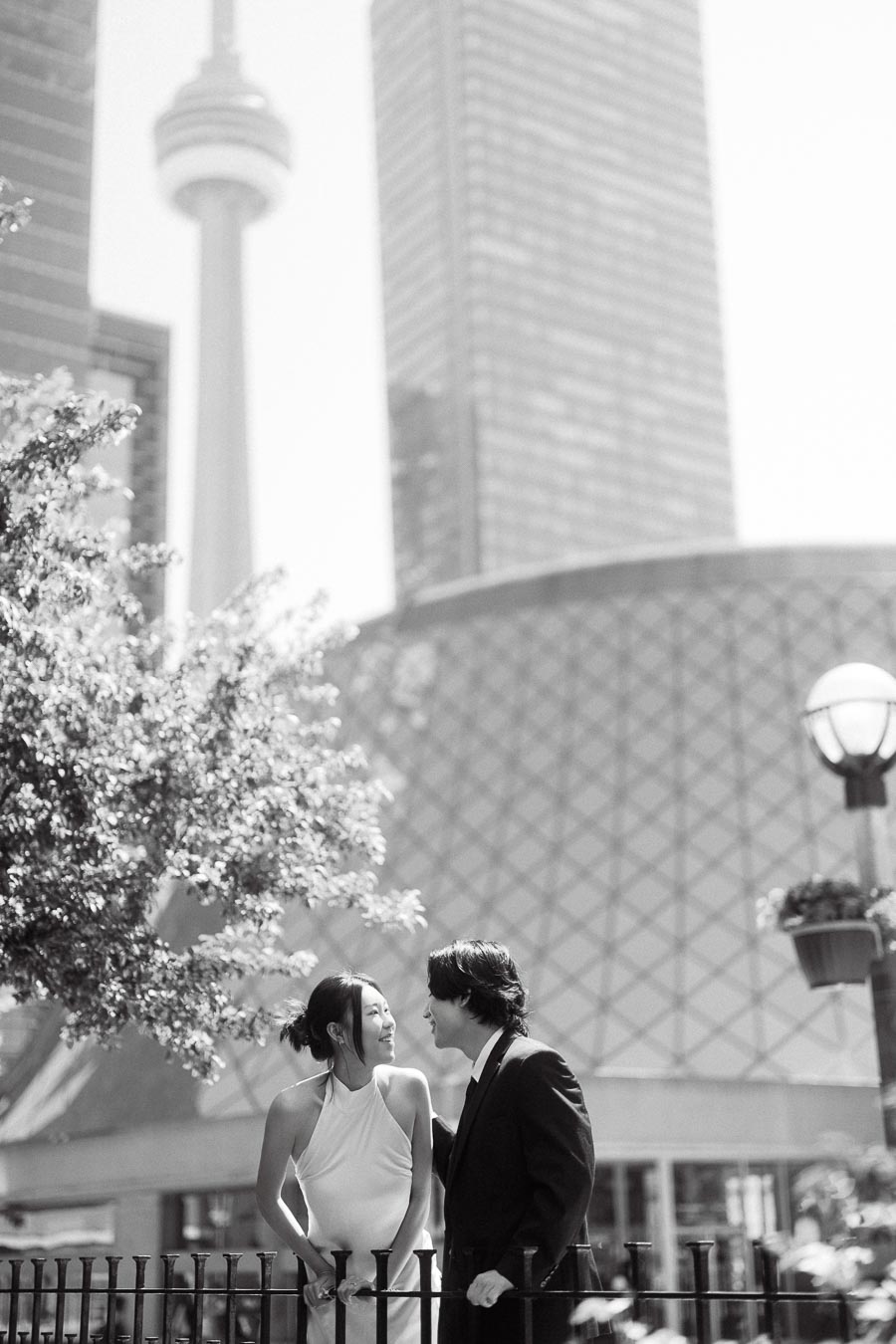 Couple smiling at each other by a fence in downtown Toronto, with the CN Tower and modern buildings in the background.