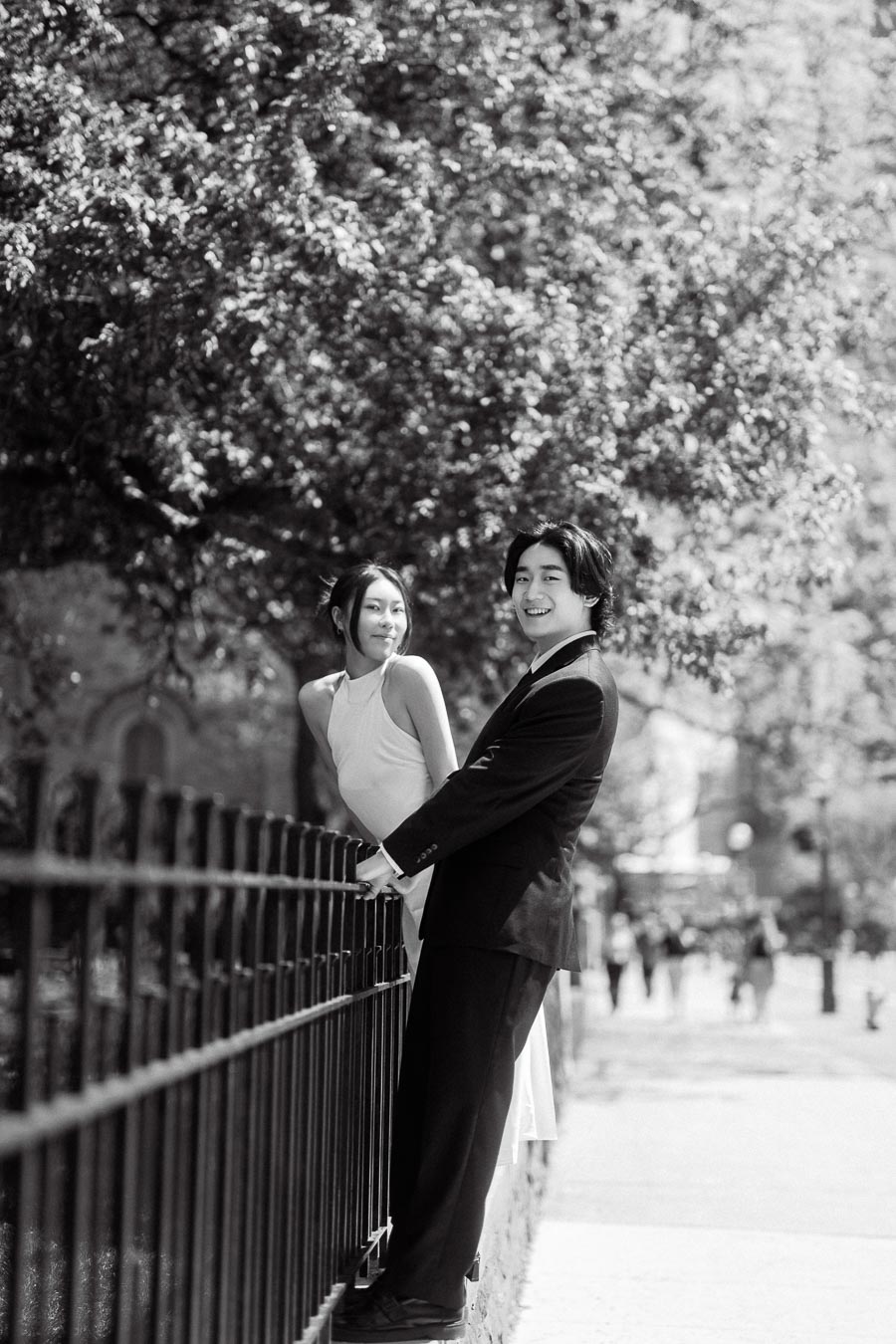 Couple dressed formally leaning on an iron fence, smiling at each other with leafy trees and city street in the background.