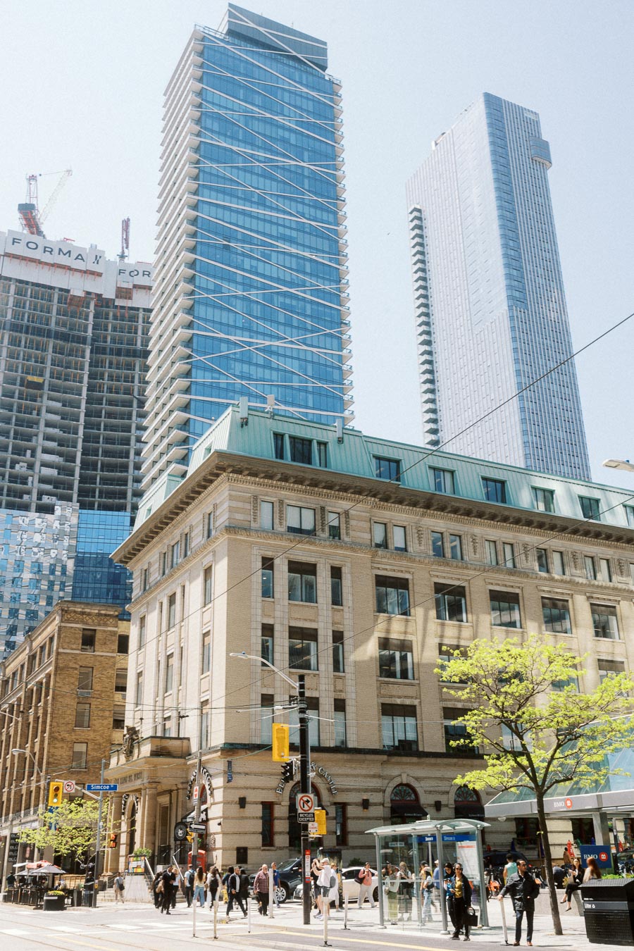 Pedestrians crossing a downtown Toronto street surrounded by modern glass skyscrapers and historic architecture on a sunny day.
