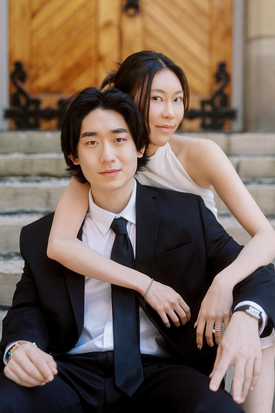 Couple sitting close together on stone steps, one in a black suit and the other in a white dress, with wooden church doors in the background.