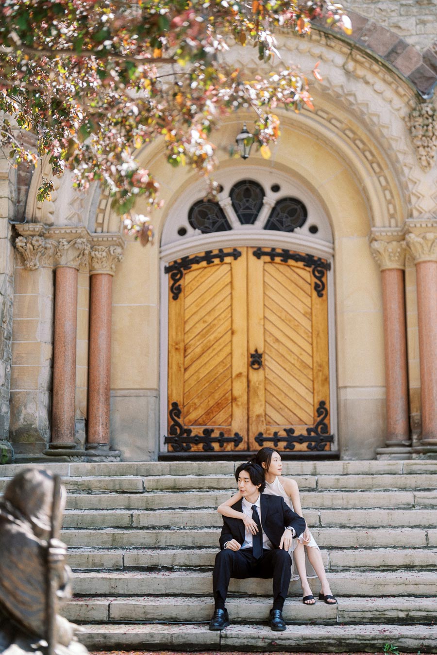 Couple sits on stone steps in front of ornate wooden church doors, with tree branches above and sunlight filtering through.