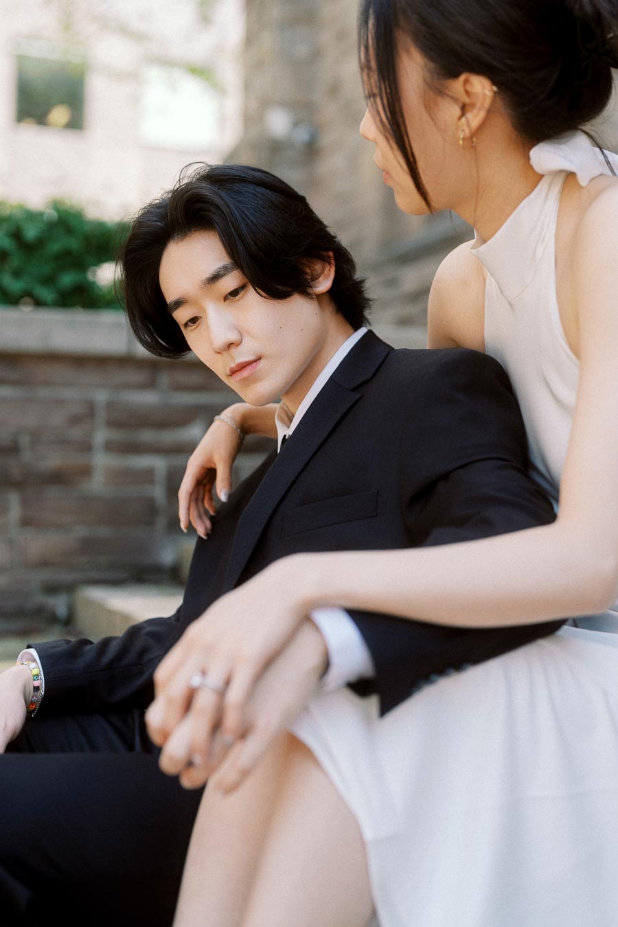 Jun and Alex sitting closely on stone steps, Alex's arm around Jun, both in formal attire, sharing an intimate moment outdoors.