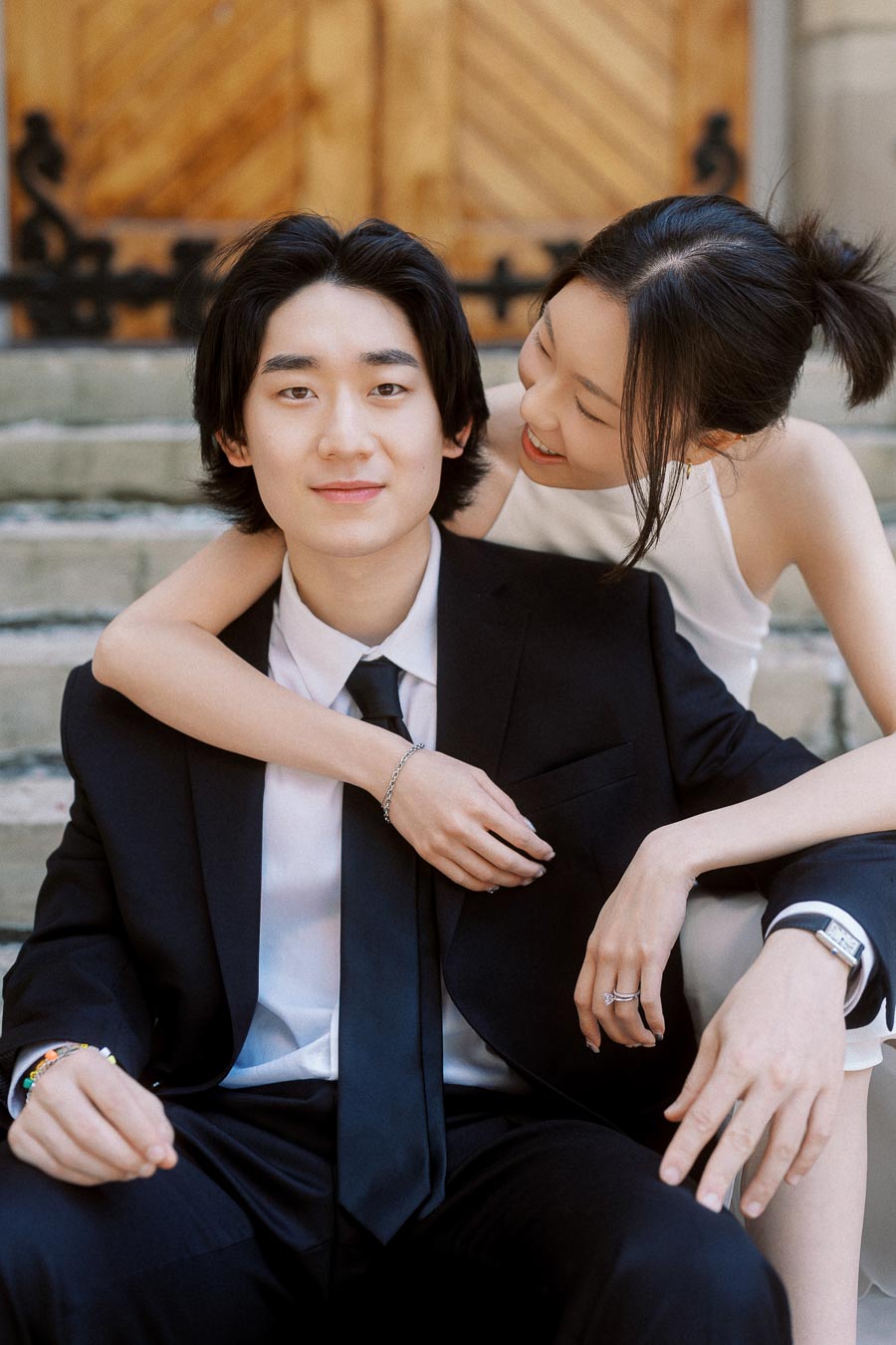 Couple sitting on church steps, one in a black suit and the other in a white dress, sharing a joyful embrace.