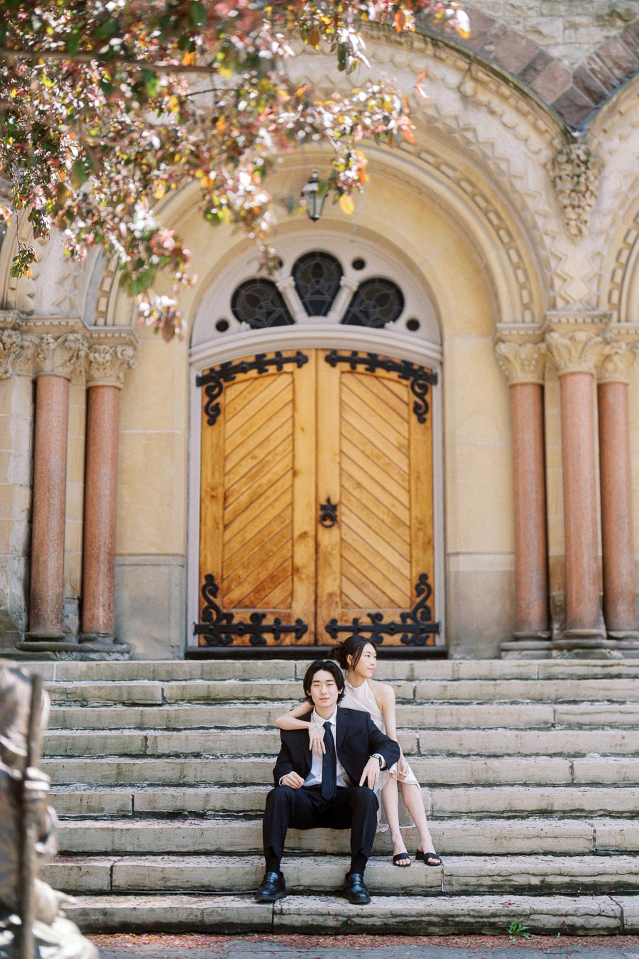 Couple seated on stone steps in front of ornate wooden church doors, surrounded by columns and leafy branches, dressed in formal attire.
