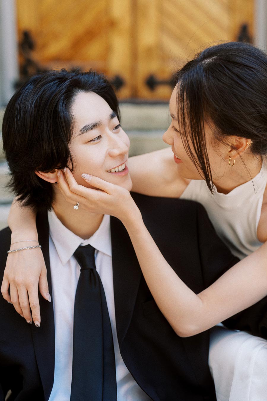Couple smiling and embracing closely on stone steps in front of wooden church doors, both dressed elegantly for an engagement photoshoot.