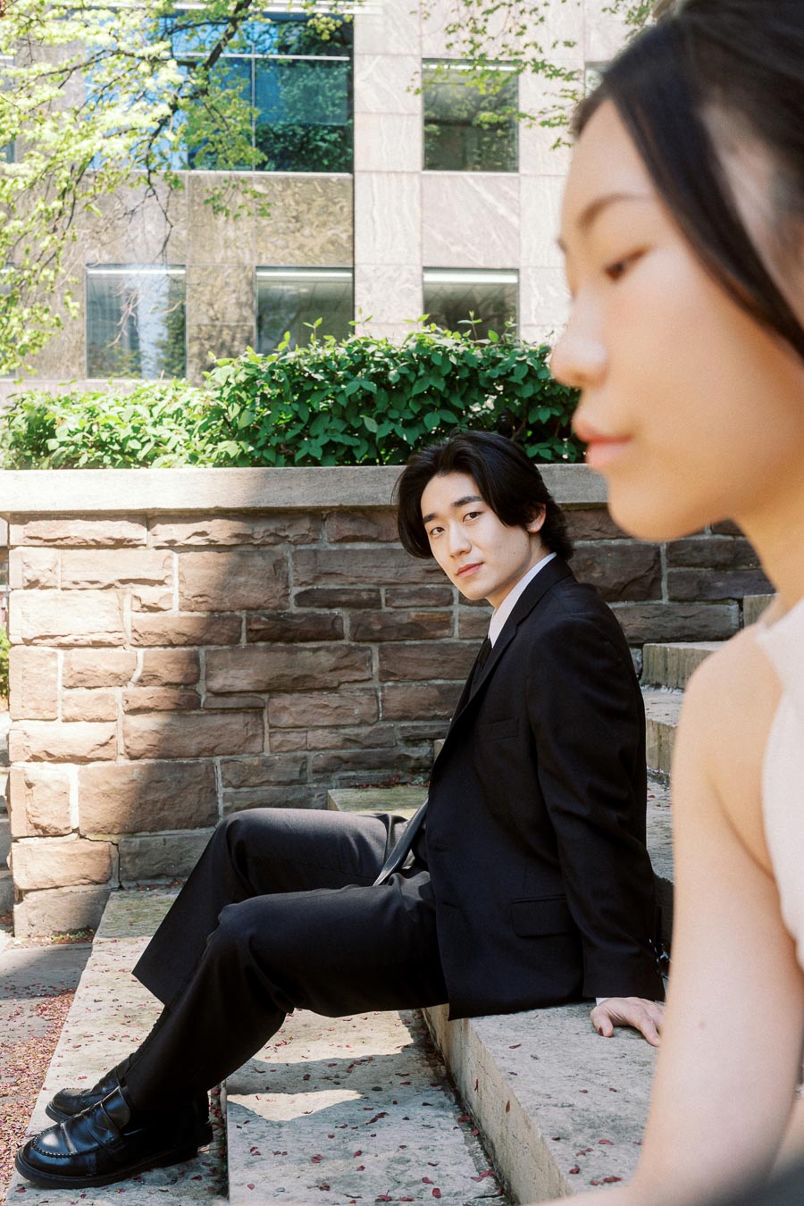 Person in a black suit sits on stone steps, looking at the camera, while another person in white is softly blurred in the foreground, with urban greenery and stone wall behind.