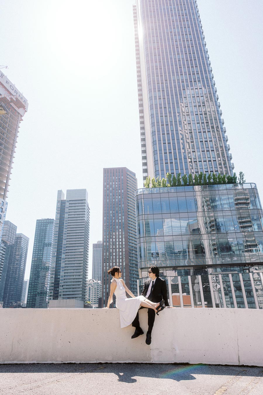 Couple sitting on a rooftop ledge in downtown Toronto, surrounded by tall glass buildings and bright sunlight, wearing formal engagement outfits.
