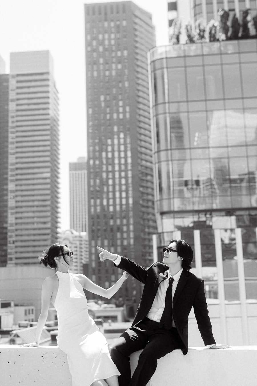 Couple in formal attire and sunglasses sitting on a rooftop ledge, smiling and interacting with downtown Toronto skyscrapers in the background.