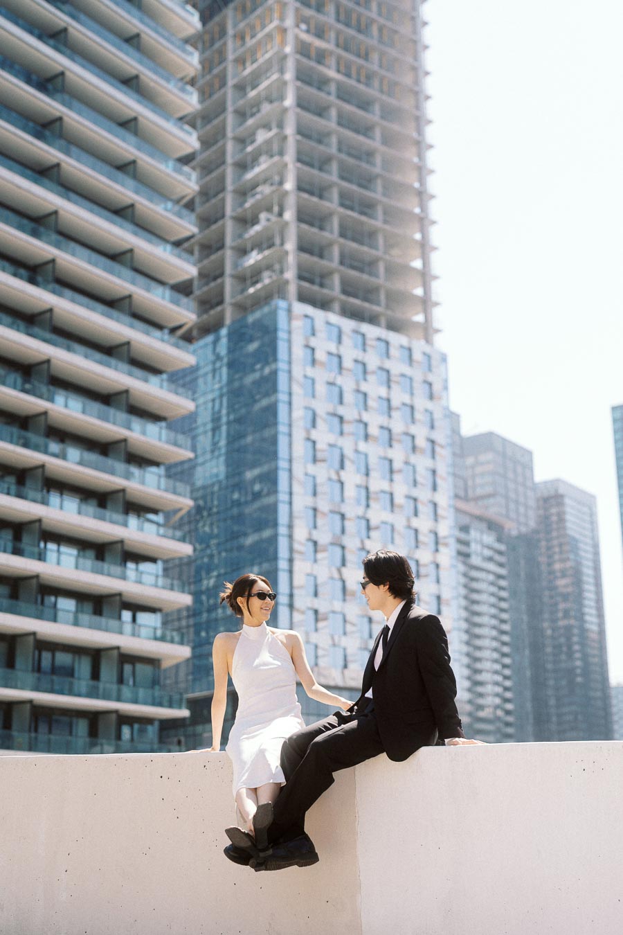 Couple sitting together on a rooftop ledge, smiling at each other, surrounded by tall modern buildings in downtown Toronto.