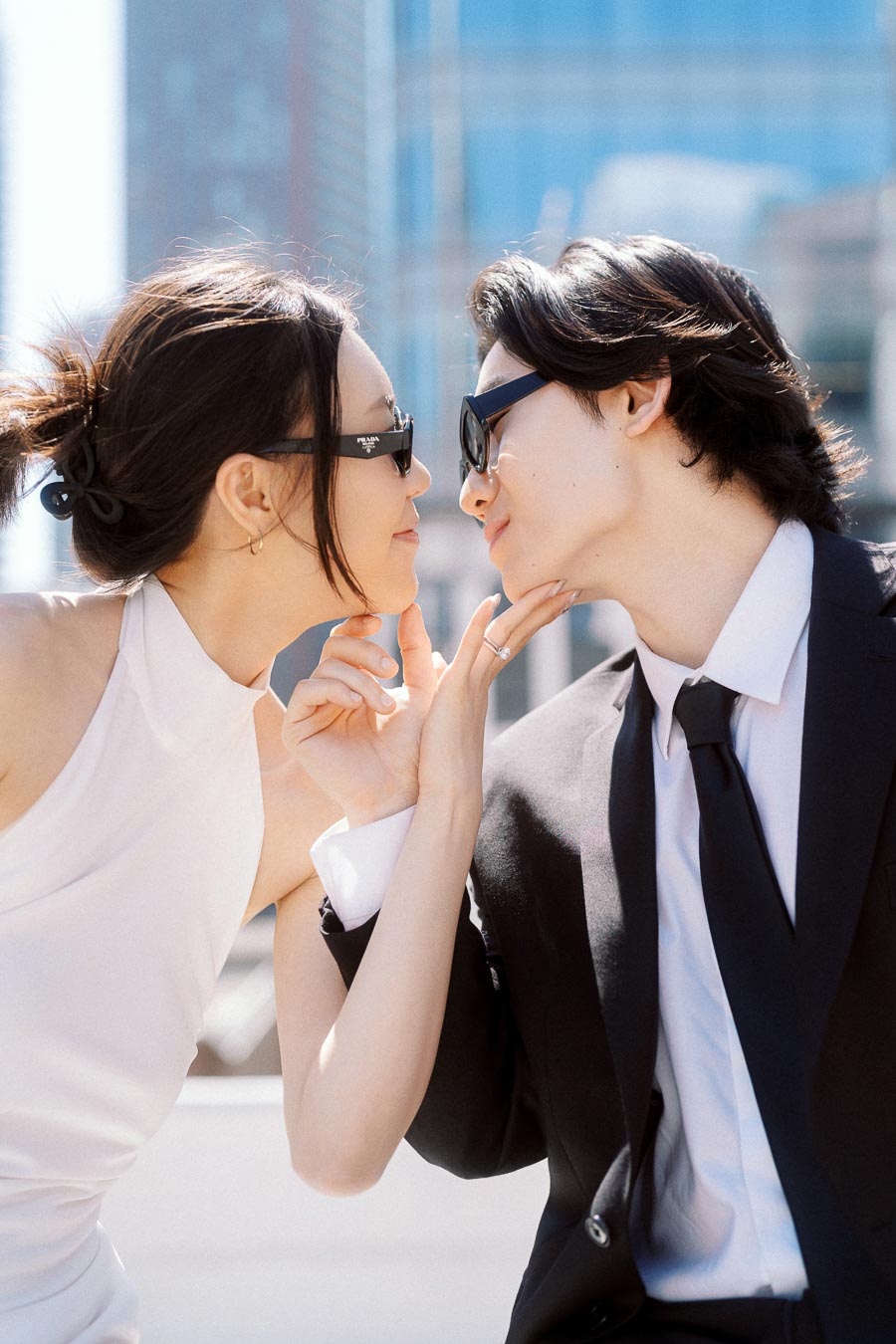 Couple in sunglasses leaning in for a kiss on a rooftop with city buildings behind them.