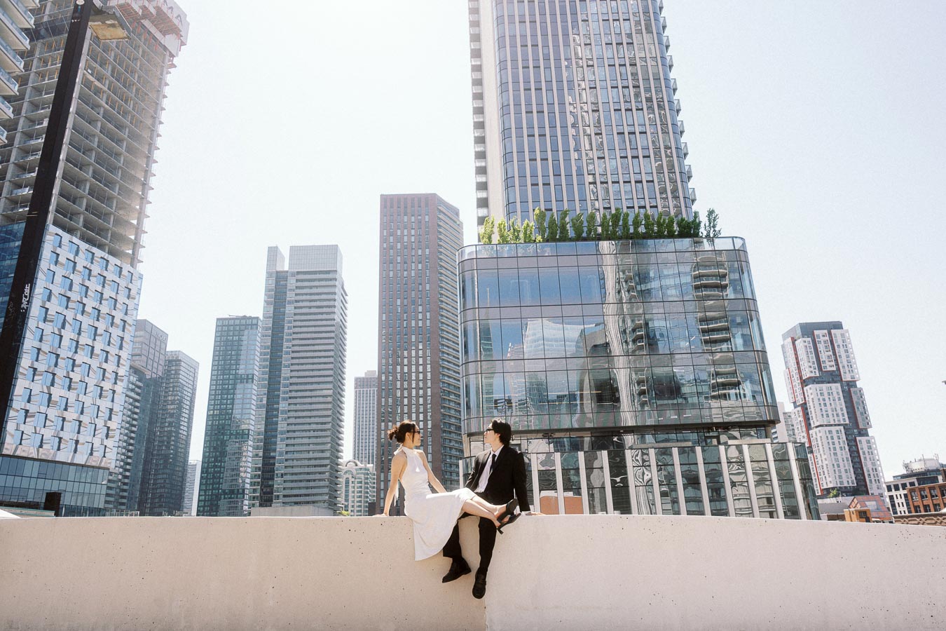 Couple sitting on a rooftop ledge in downtown Toronto, with modern glass skyscrapers and city skyline in the background.