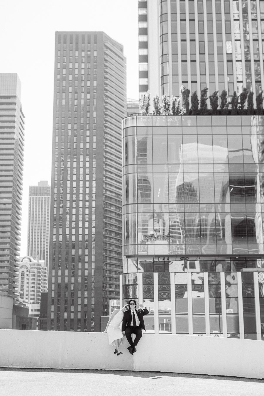 Couple sitting side by side on a rooftop ledge, surrounded by tall glass and concrete buildings in downtown Toronto, both dressed formally.