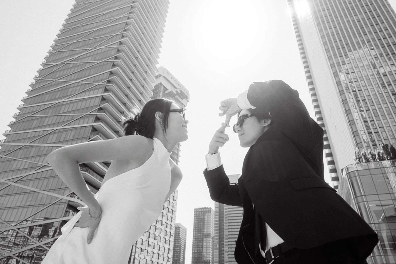 Two people in formal outfits and sunglasses playfully posing together among tall glass buildings in downtown Toronto, sunlight streaming overhead.