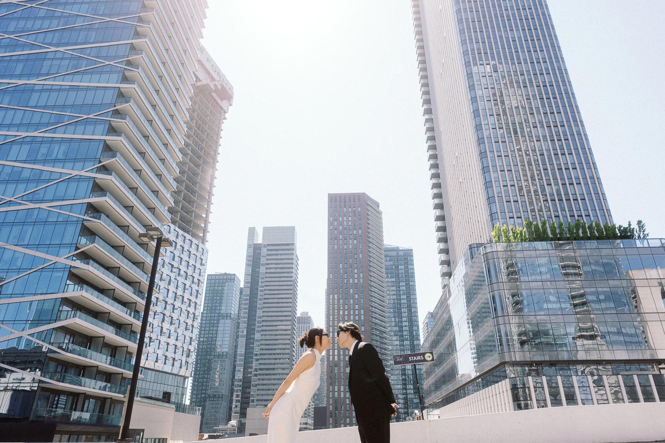 Two people, one in a white dress and one in a black suit, leaning in for a kiss on a rooftop with tall glass skyscrapers in downtown Toronto.