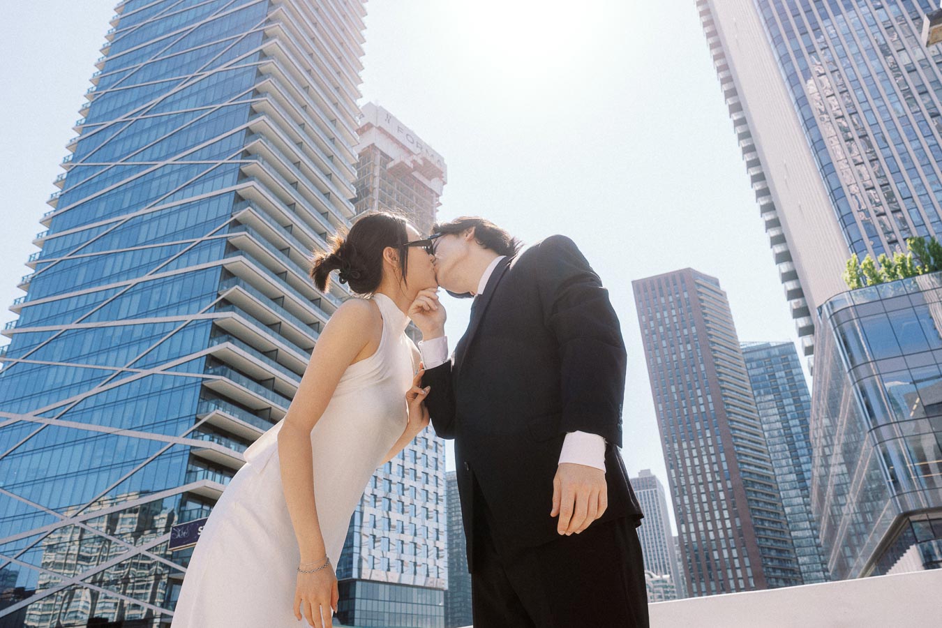 A couple dressed in a white dress and black suit kiss under bright sunlight, framed by tall glass skyscrapers in downtown Toronto.