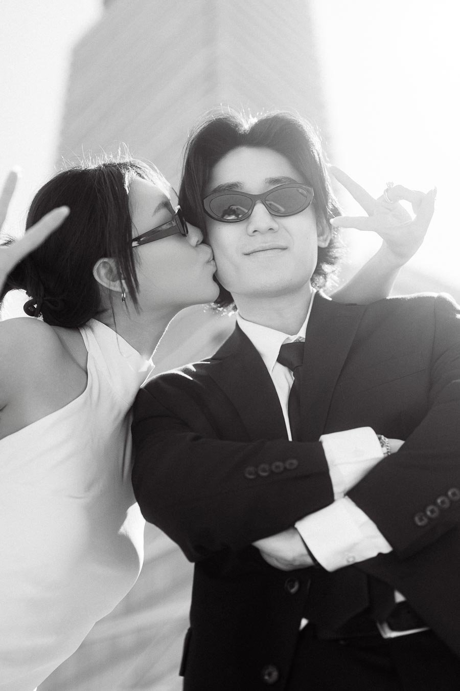 Jun kisses Alex's cheek while both wear sunglasses and pose playfully on a sunny rooftop with a city building behind them.