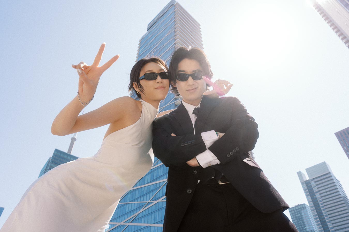 Couple in formal attire and sunglasses posing playfully with peace signs in front of downtown Toronto skyscrapers on a sunny day.