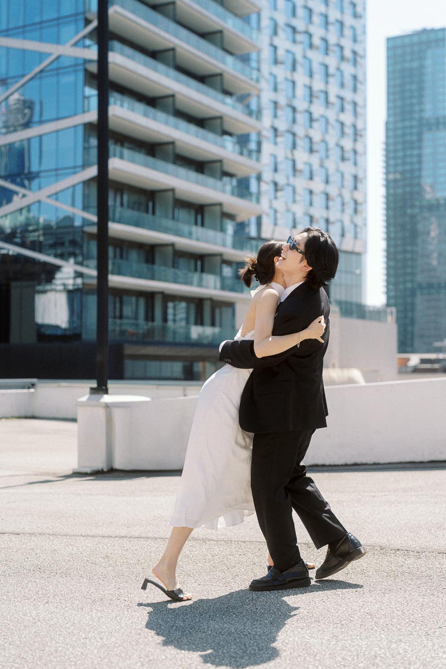 Couple embracing and smiling on a rooftop with modern glass buildings in downtown Toronto, both wearing sunglasses and stylish outfits.